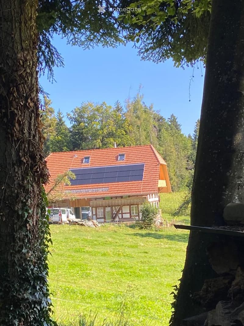 Two-story house with red-tiled roof, large windows, and solar panels on the roof, surrounded by a green lawn and trees.