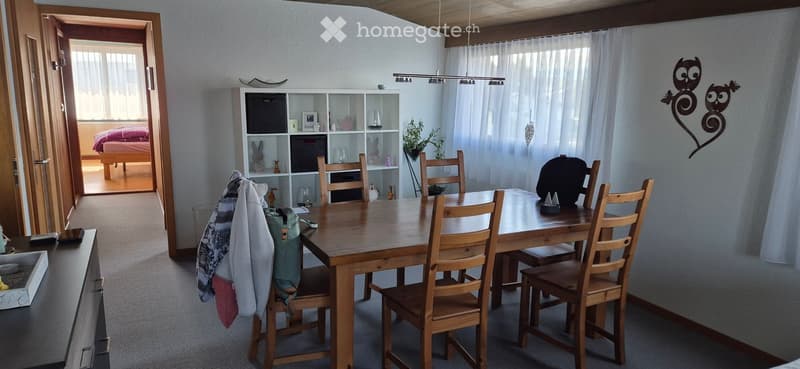 Bright dining area with a wooden dining table and chairs, a white shelf with decorative items, and a view into a bedroom with a bed and pink bedding.