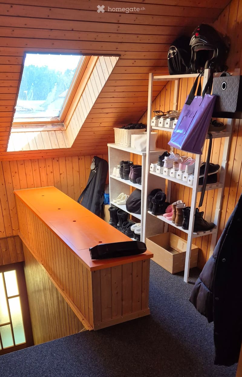 A small, cozy room with wooden walls and a slanted ceiling, featuring a long wooden bench, a white shelf filled with shoes and bags, and a window offering a scenic view.