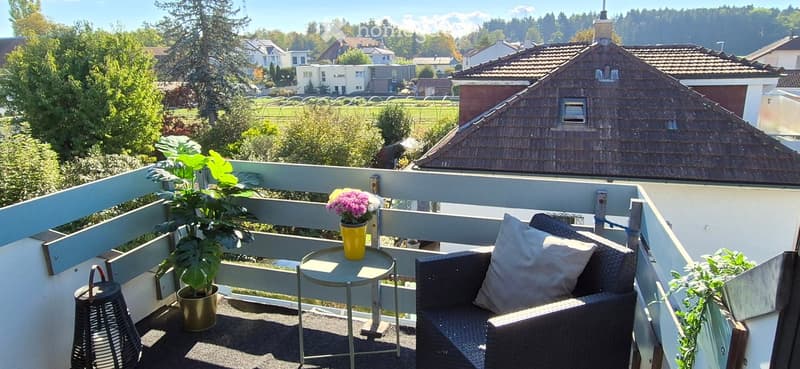 Balcony with a view of a suburban neighborhood, featuring a small sofa, a round table with a flower pot, and a handbag.