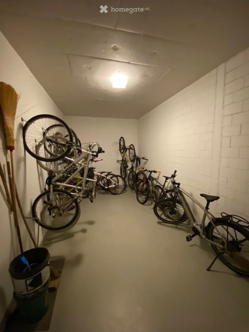 A narrow, well-lit hallway with white brick walls and a concrete floor. Several bicycles are stored on racks along the walls, and a broom and dustpan are leaning against the wall on the left side.