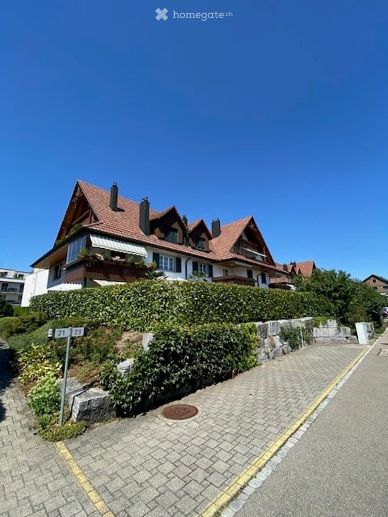 A two-story house with a red-tiled roof, white walls, and multiple windows. The house features a balcony and is surrounded by a well-maintained hedge and garden. The entrance is marked by a stone wall and a paved pathway.