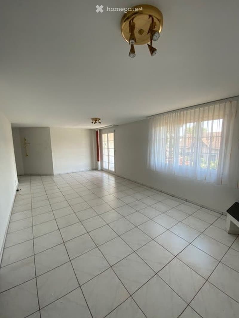 Empty room with white tiled flooring, white walls, large window with white curtains, and a simple door. No furniture or equipment present.