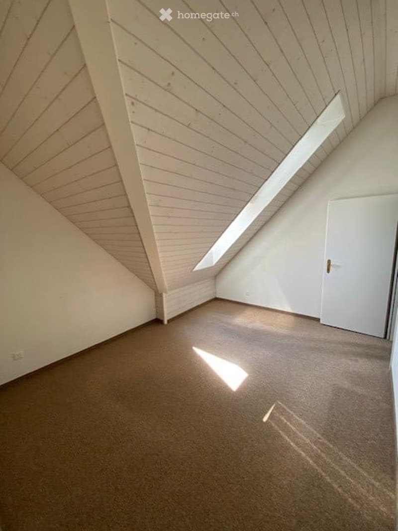 Empty attic room with wooden slanted ceilings, carpeted floor, white walls, and a skylight window letting in natural light.