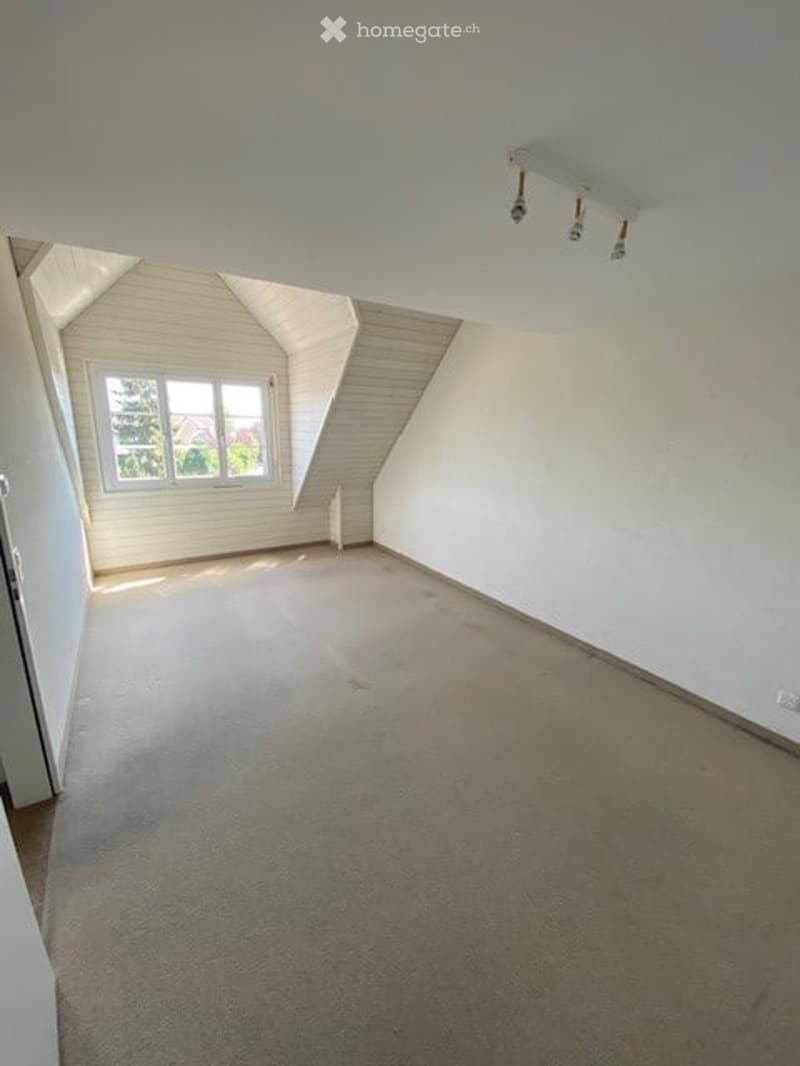Empty attic room with sloped ceilings, white walls, carpeted floor, and a single window letting in natural light.