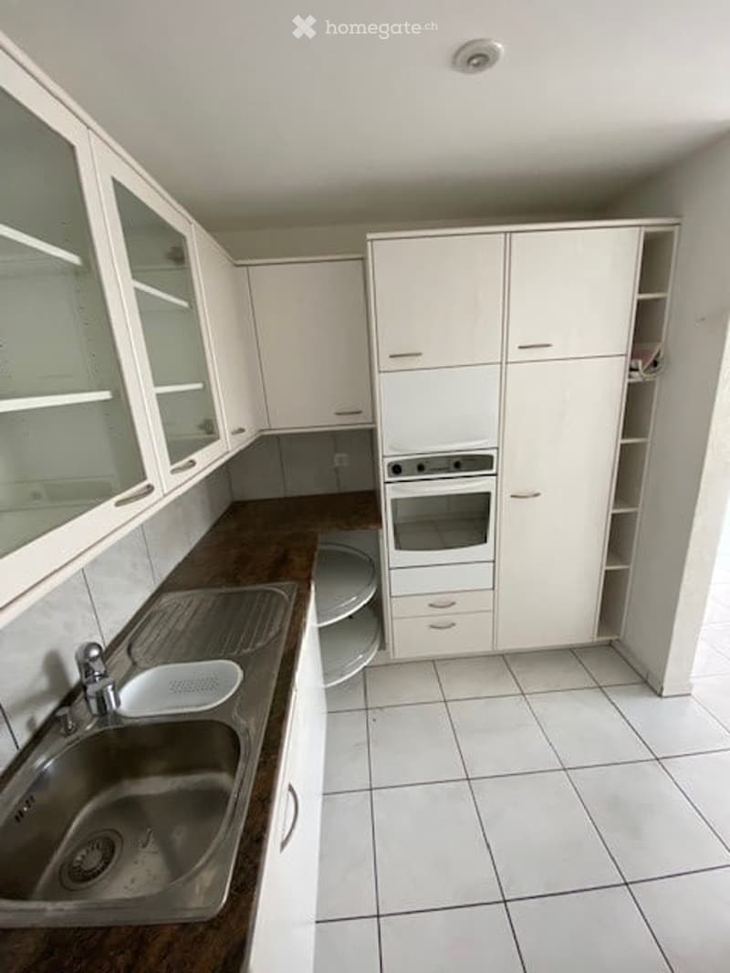Modern kitchen with white cabinets, stainless steel sink, and built-in oven. The countertop is dark brown, and the floor is tiled.
