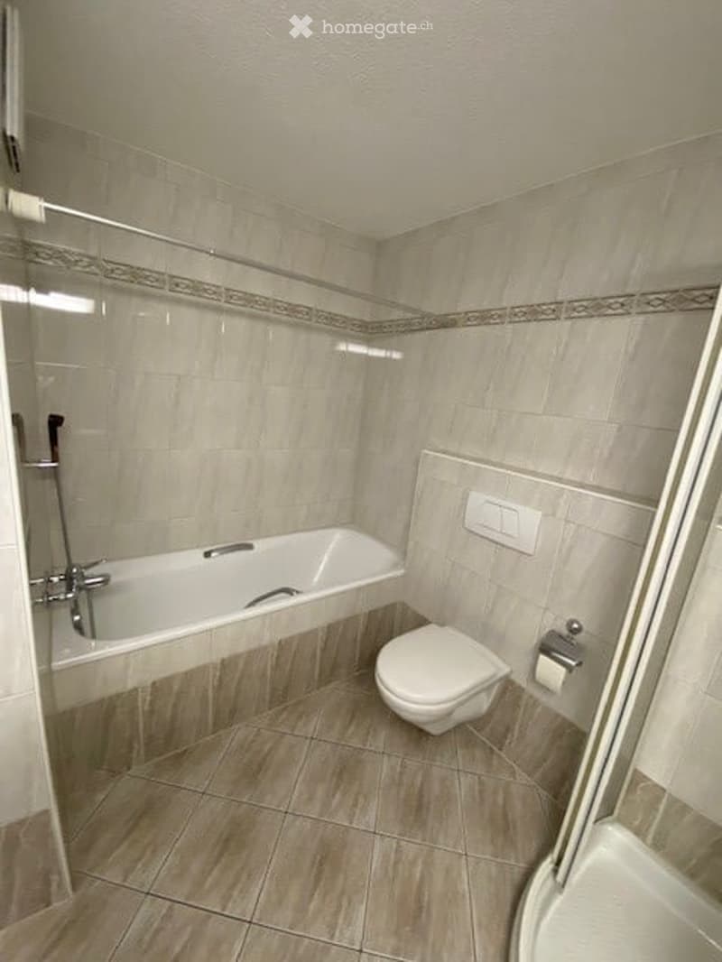 Modern bathroom with a bathtub, toilet, and shower area. The walls are tiled in light beige with a decorative border, and the floor is tiled in a warm wood tone.