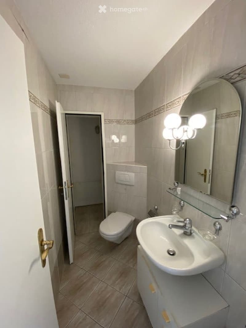 Modern bathroom with a white sink, toilet, large mirror, and wall-mounted light fixture. The walls are tiled with a decorative border, and the floor is tiled in a warm wood tone.