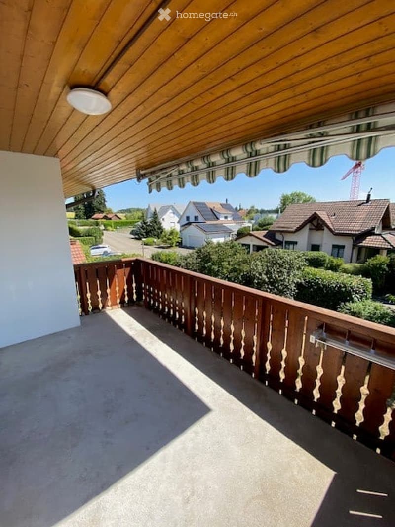 Wooden balcony with a railing, covered by a wooden awning, overlooking a residential area with houses and greenery.