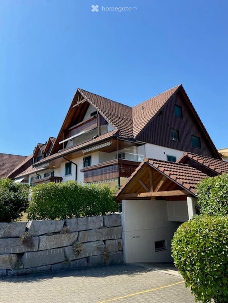 Two-story house with a red-tiled roof, wooden facade, and a garage entrance. The house features balconies and is surrounded by greenery.