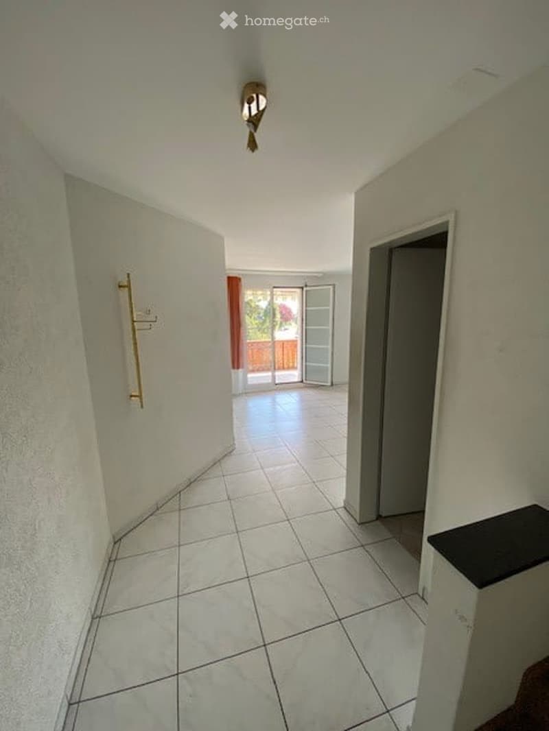 Empty hallway with white tiled flooring, white walls, and a ceiling light fixture. A door leads to a balcony or terrace area with a view of outdoor space.