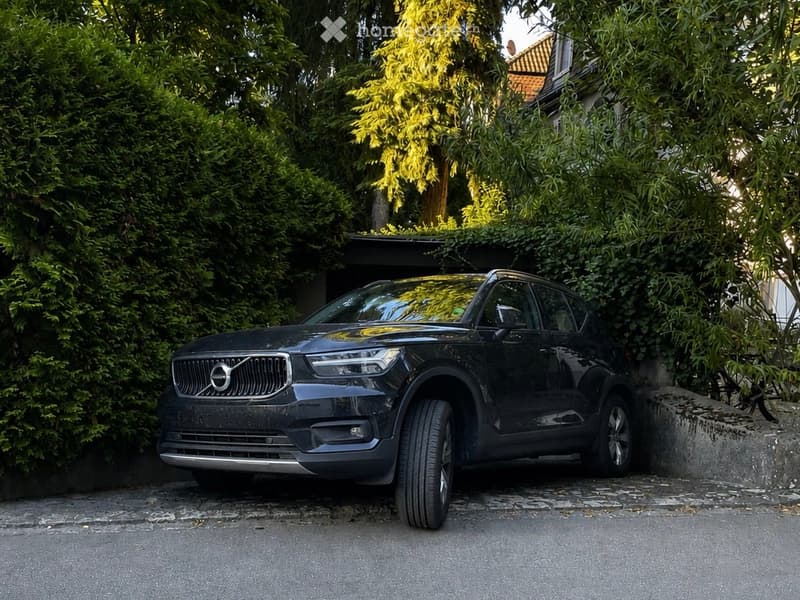 A black Volvo XC60 SUV parked in a driveway next to a house with lush greenery and a stone wall.