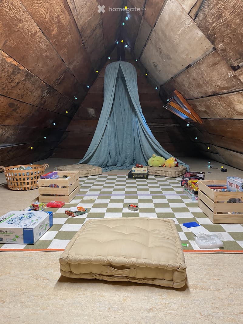 A cozy attic space with wooden beams, a large window, and a blue curtain creating a tent-like area. The room features a checkered floor, a large cushioned sofa, wooden crates, a wicker basket, and various toys and books scattered around.