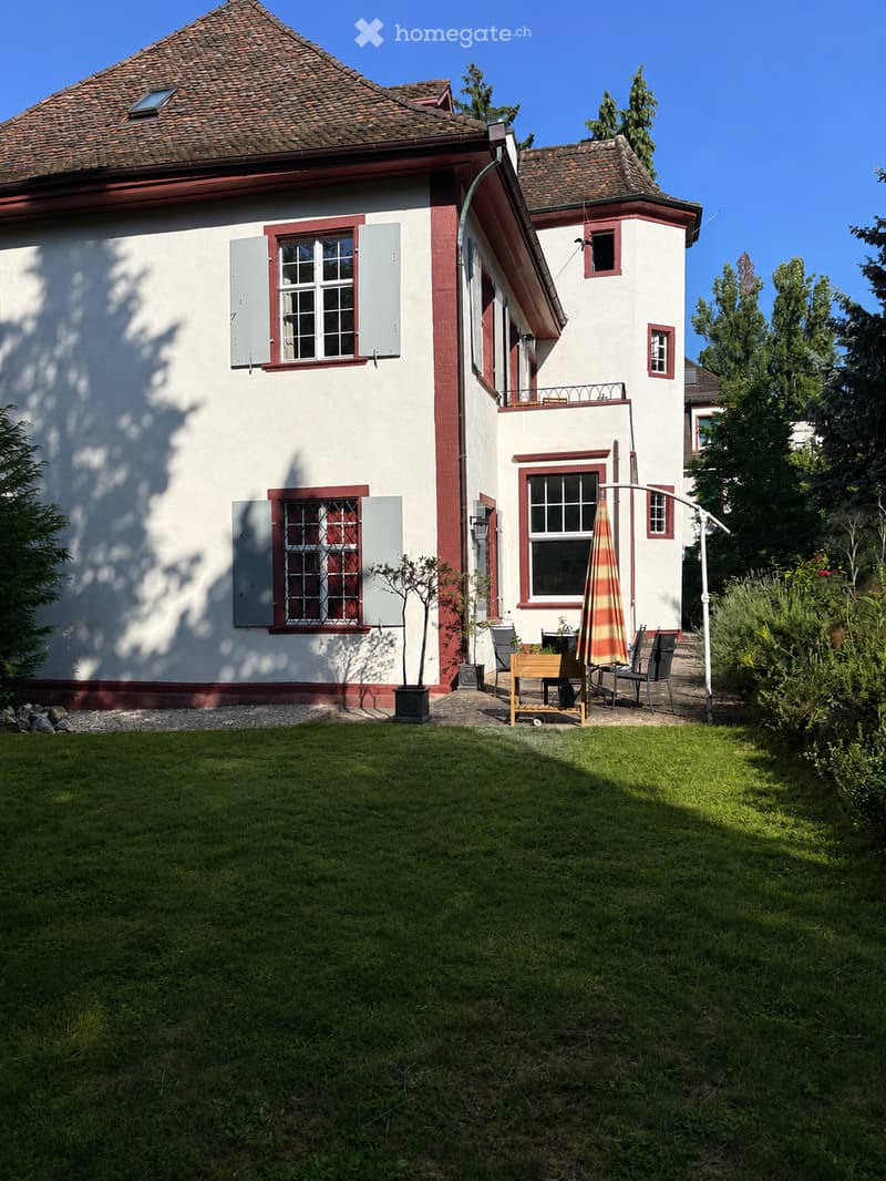 A two-story house with white walls, red trim, and a tiled roof. The front yard features a well-maintained lawn, a small patio area with outdoor furniture, and a small tree in a planter. The house has multiple windows with shutters and a small balcony on t
