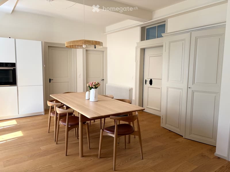 Modern dining area with a wooden dining table, four chairs, a vase with flowers, and built-in white cabinets with an oven.