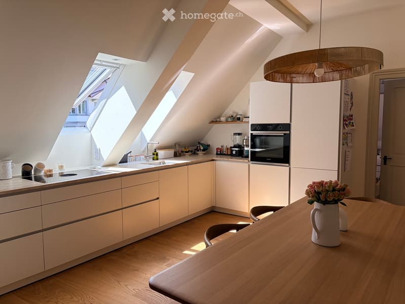 Bright, modern kitchen with light-colored cabinets, wooden flooring, and skylight windows. Includes a sink, oven, blender, table, and vase with flowers.