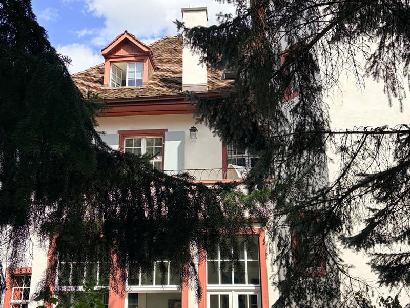 Two-story house with a tiled roof, white walls, red trim, and multiple windows partially obscured by tree branches.