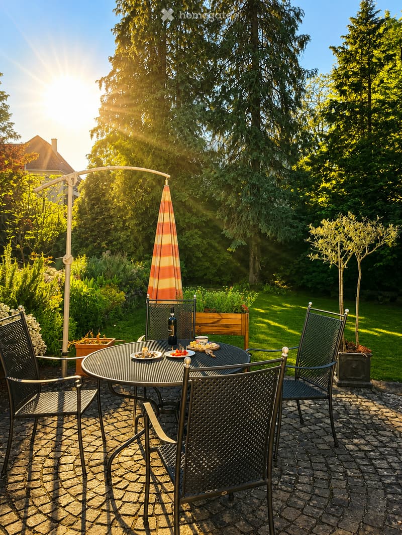 A sunny outdoor patio with a round metal table, four black metal chairs, an umbrella, and a bottle on the table. The area is surrounded by lush greenery and trees.
