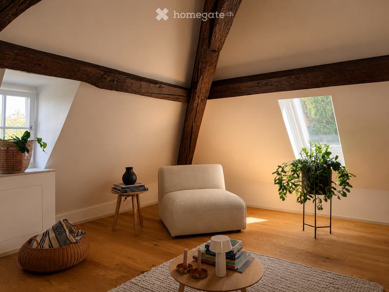 Cozy attic room with wooden beams, large windows, a cream-colored sofa, a small wooden table with books and a lamp, and decorative plants.