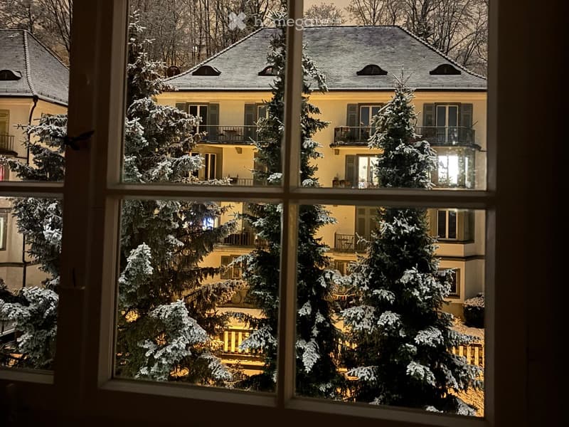View from a window of a snow-covered residential building with illuminated windows and a well-lit garden area.
