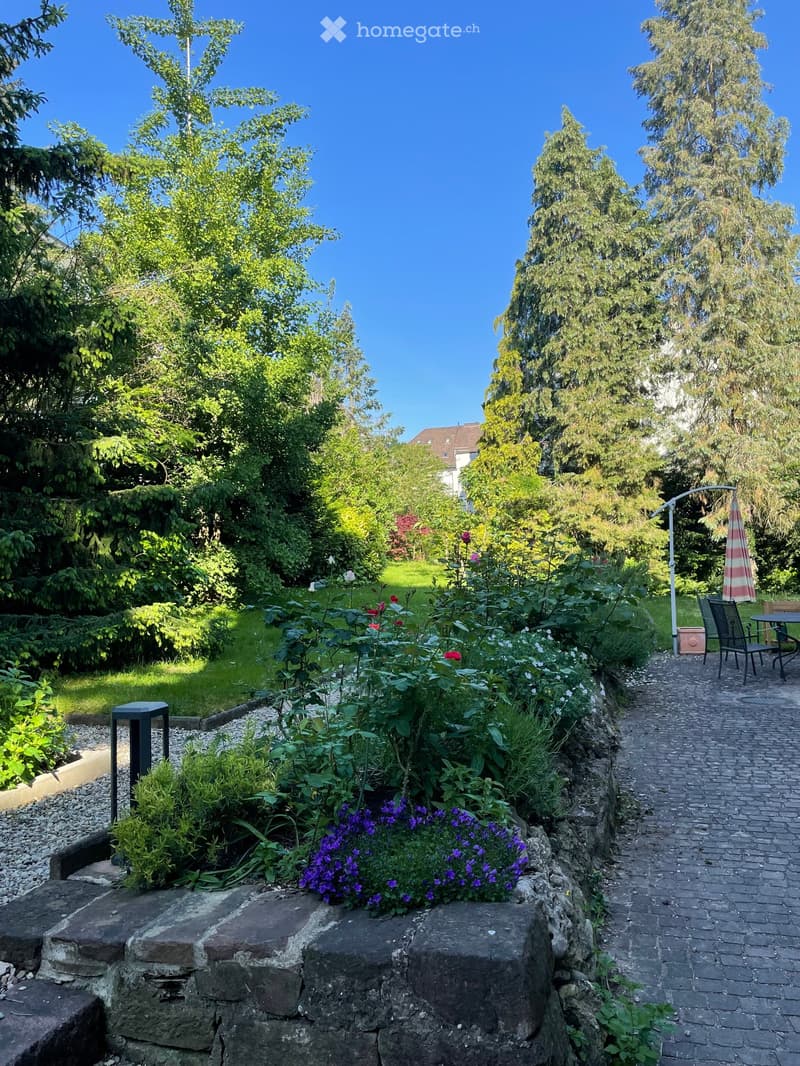 A well-maintained garden with a variety of flowers, shrubs, and trees, featuring a paved pathway, outdoor seating area, and a clear blue sky.