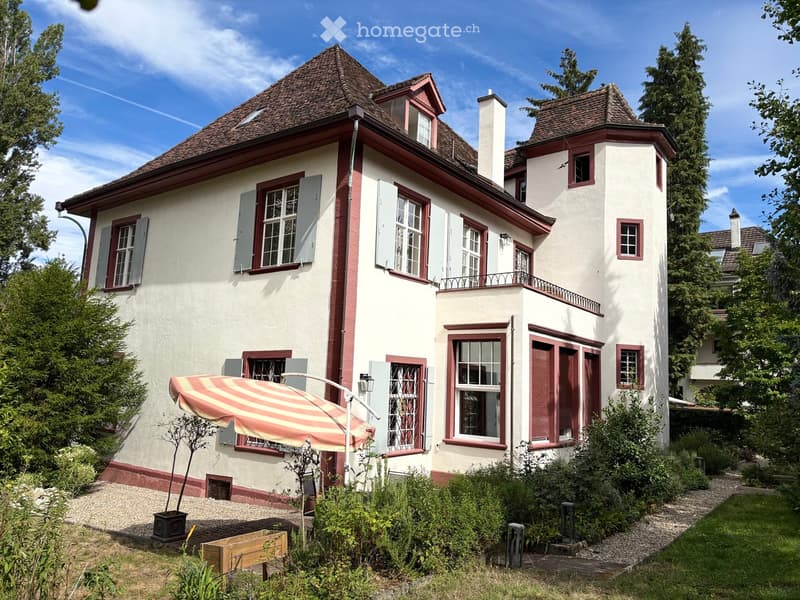 Two-story house with white walls, red trim, multiple windows, and a tiled roof. Features a small balcony, garden area with plants, and a striped umbrella near the entrance.