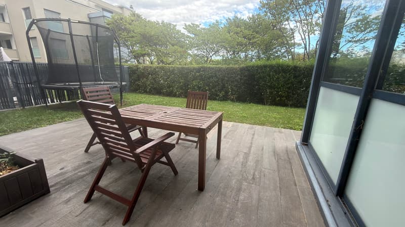 Wooden patio with a table and chairs, surrounded by a grassy area and a hedge, with a trampoline in the background.