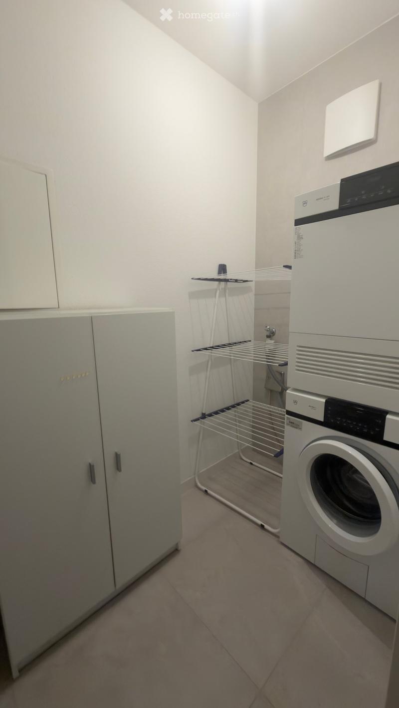 A small, well-lit room with a washing machine, drying rack, and built-in cabinets.