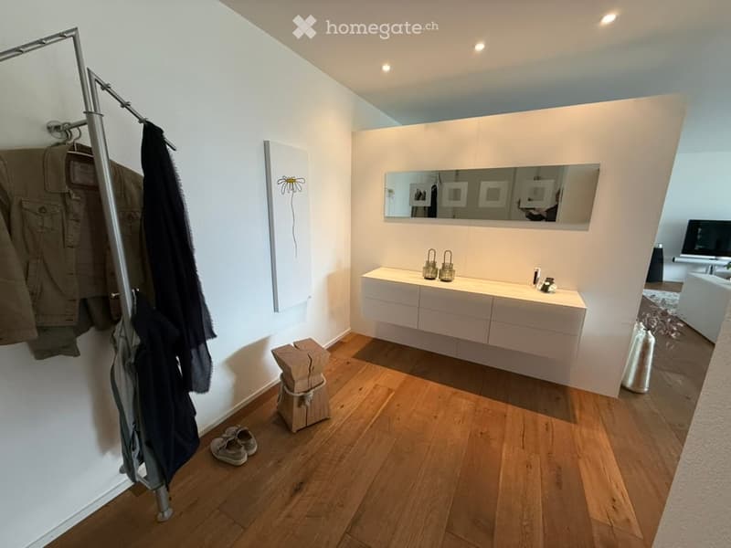 Empty room with wooden flooring, white walls, a large mirror, and a white vanity with drawers. There is a coat rack with jackets and a small wooden stool with shoes nearby.