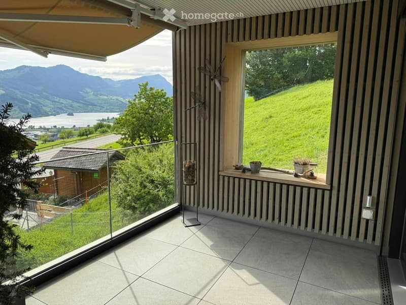 Modern balcony with large windows offering scenic mountain and lake views, tiled floor, glass railing, and decorative items on the windowsill.