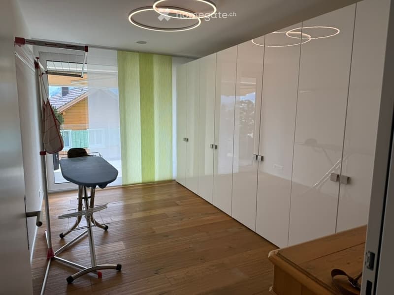 Modern, minimalist room with white built-in wardrobes, wooden flooring, and a round ceiling light fixture. An ironing board and stand are positioned near a window with green blinds.