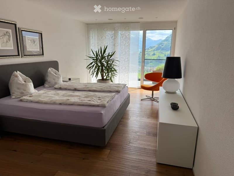 Modern bedroom with wooden flooring, large bed with white bedding, nightstand, chair, table lamp, plant, and scenic mountain view through large windows.