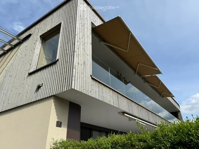 Modern two-story building with a combination of wooden and metal exterior walls, large glass windows, and a covered balcony area with retractable awnings.