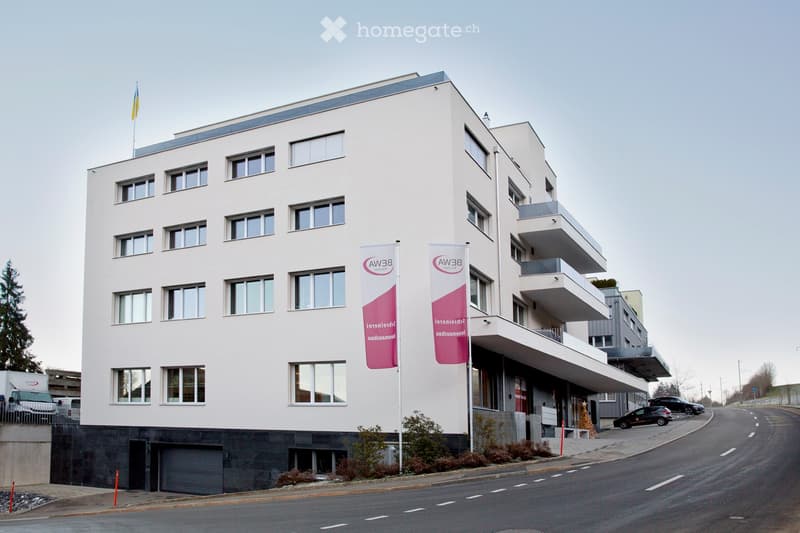 Modern multi-story building with a light-colored facade, multiple windows, and a flagpole with a flag. There are banners on the side and a small landscaped area in front.