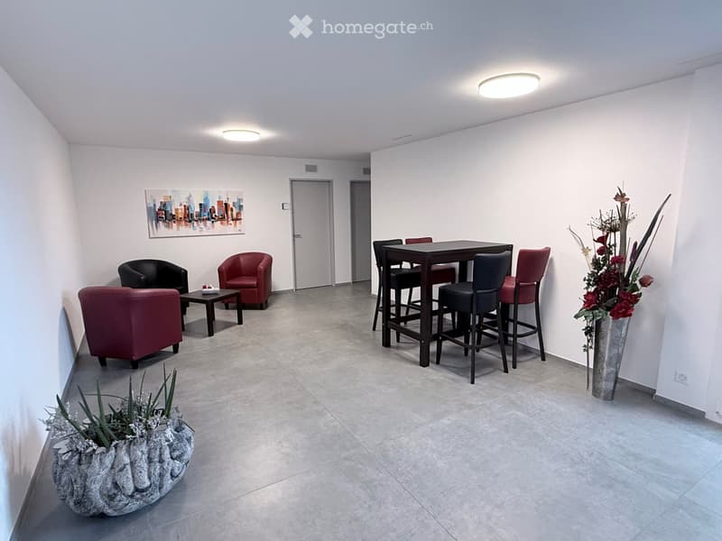 Modern, well-lit room with a mix of seating and a small dining area. Features include a red sofa, black armchair, small coffee table, bar table with stools, and decorative vases.