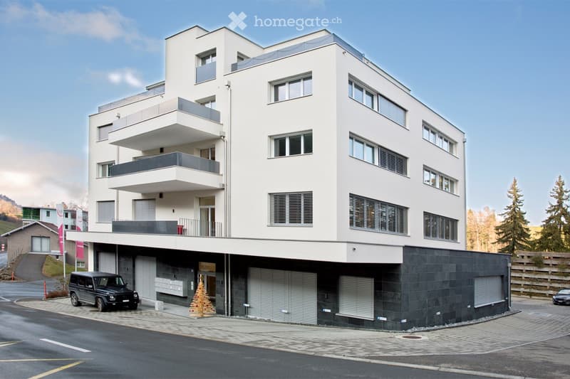 Modern multi-story building with white facade, multiple balconies, and a parking area with a black SUV parked in front.