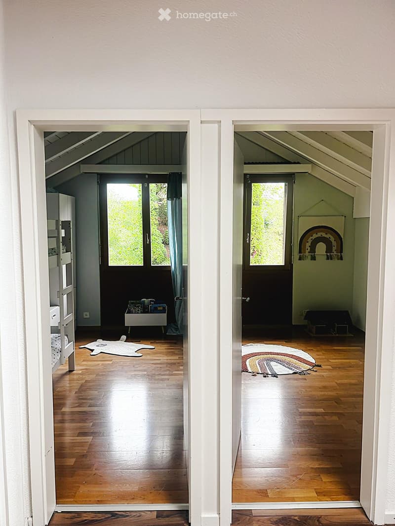 A bright, open hallway with wooden flooring and white walls, leading to two adjacent rooms with large windows and skylights, featuring simple decor and natural light.