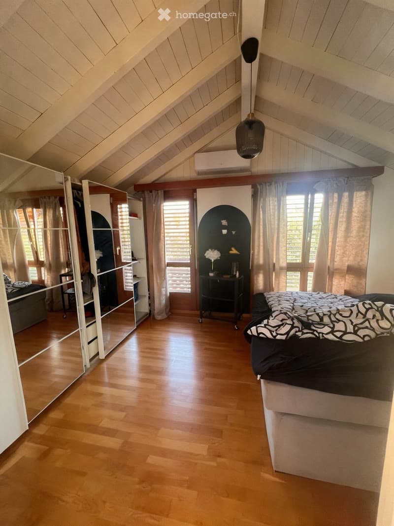 Bright attic bedroom with wooden flooring, slanted wooden ceiling, large windows with curtains, a bed with black and white bedding, a wardrobe, and a small table with a lamp.