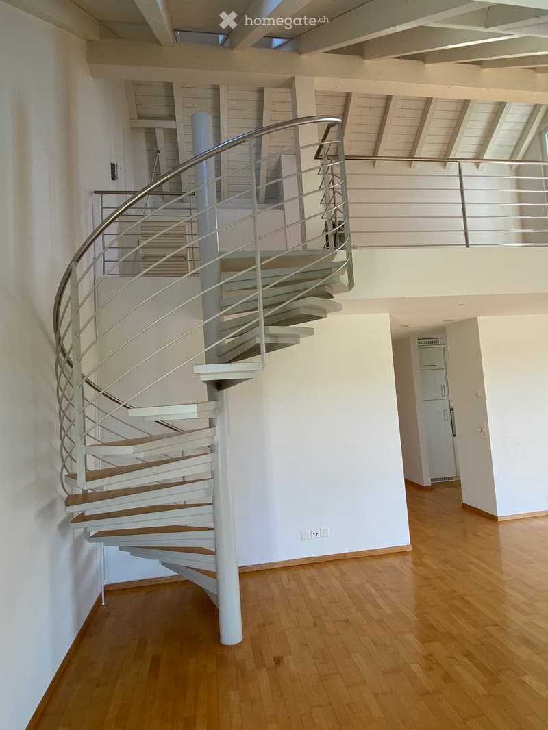 Spiral staircase with wooden steps and white railings, leading to an upper level with a glass balcony railing. The hallway has light wooden flooring and white walls.