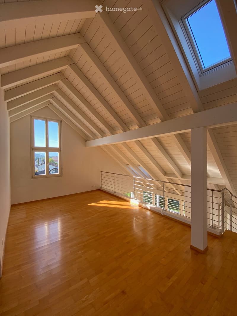 Bright, empty attic space with wooden floors, white walls, exposed wooden beams, and large windows offering natural light and views.