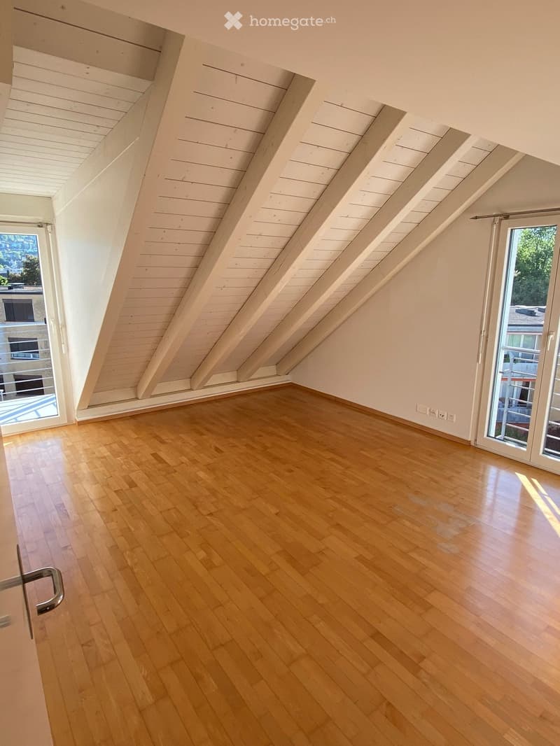 Empty attic room with wooden floors, white walls, and exposed wooden beams on the ceiling. Features large windows and sliding glass doors leading to a balcony.
