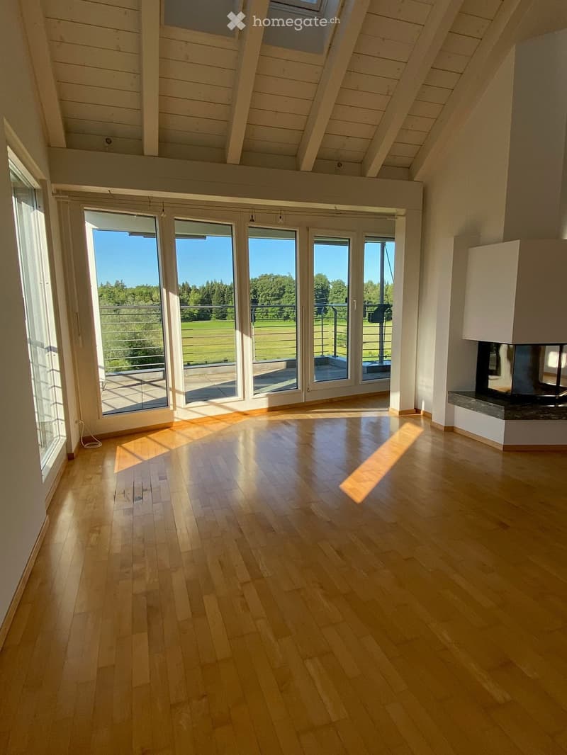 Bright, empty room with wooden flooring, large windows, and a fireplace. The room features a balcony with a view of a green landscape.