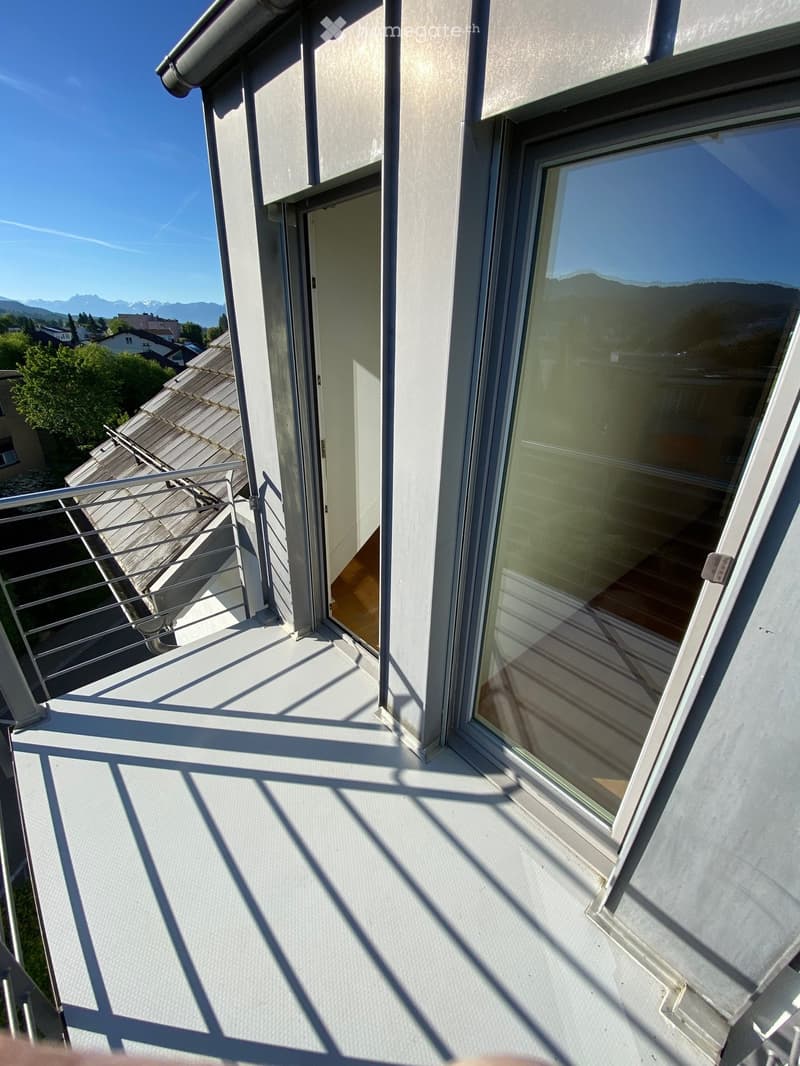 Balcony with metal railing, large window, and view of the surrounding area and mountains.