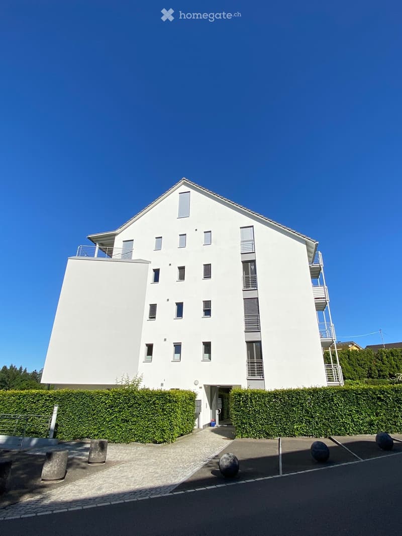 Modern two-story white building with multiple windows and balconies, surrounded by a neatly trimmed hedge and decorative spherical elements.