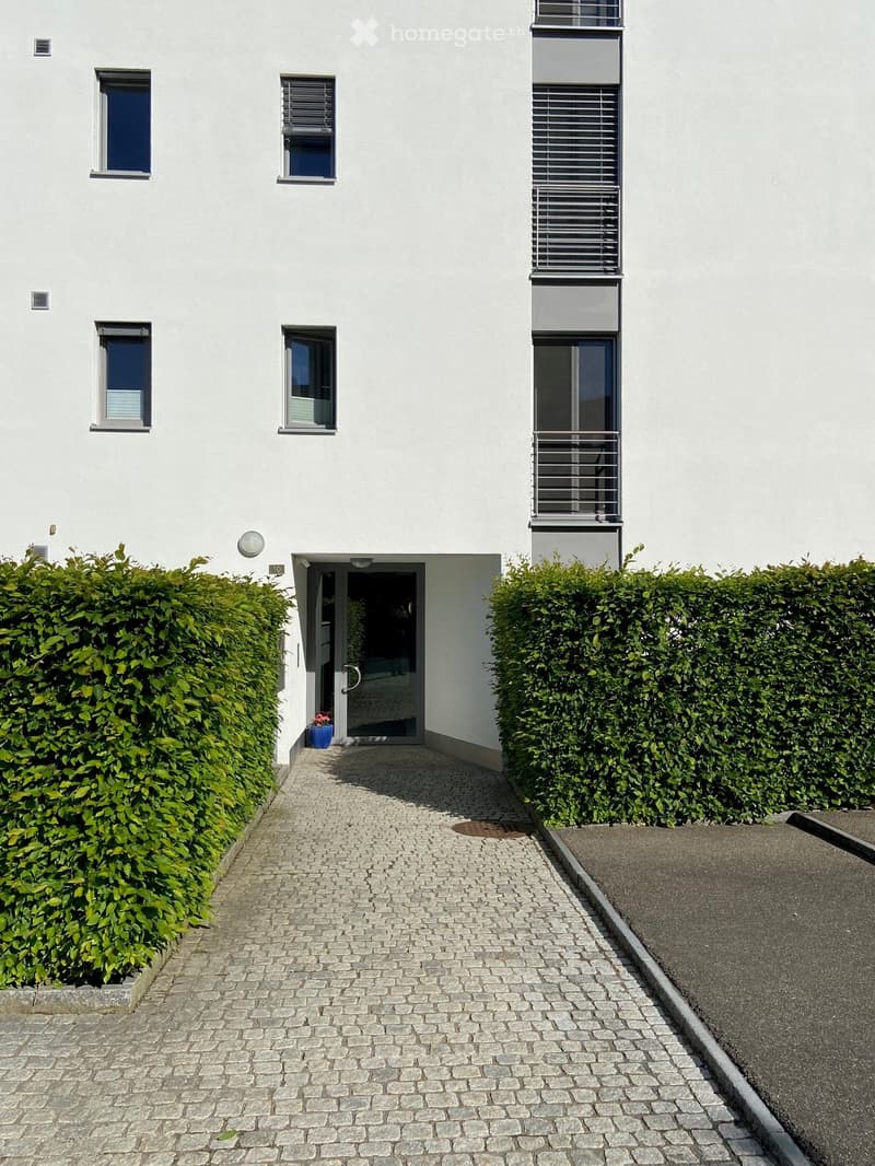 Modern two-story building with white facade, multiple windows, and a small balcony on the upper floor. The entrance features a glass door with a small potted plant nearby.
