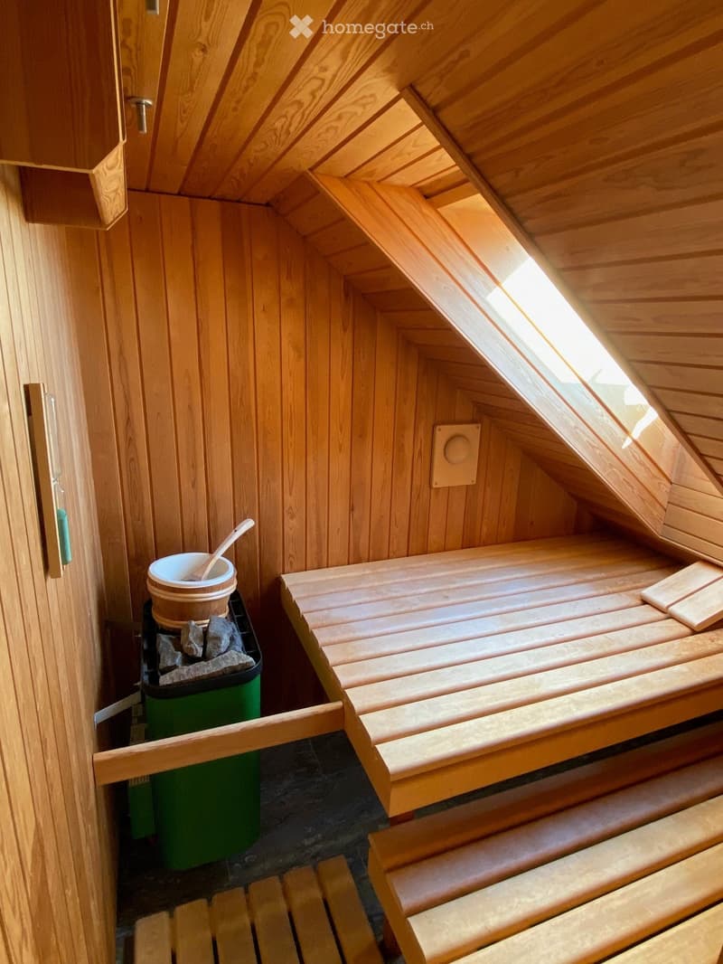 Wooden sauna interior with benches, a bucket, and a wooden spoon, featuring a skylight window and a stone-filled container.