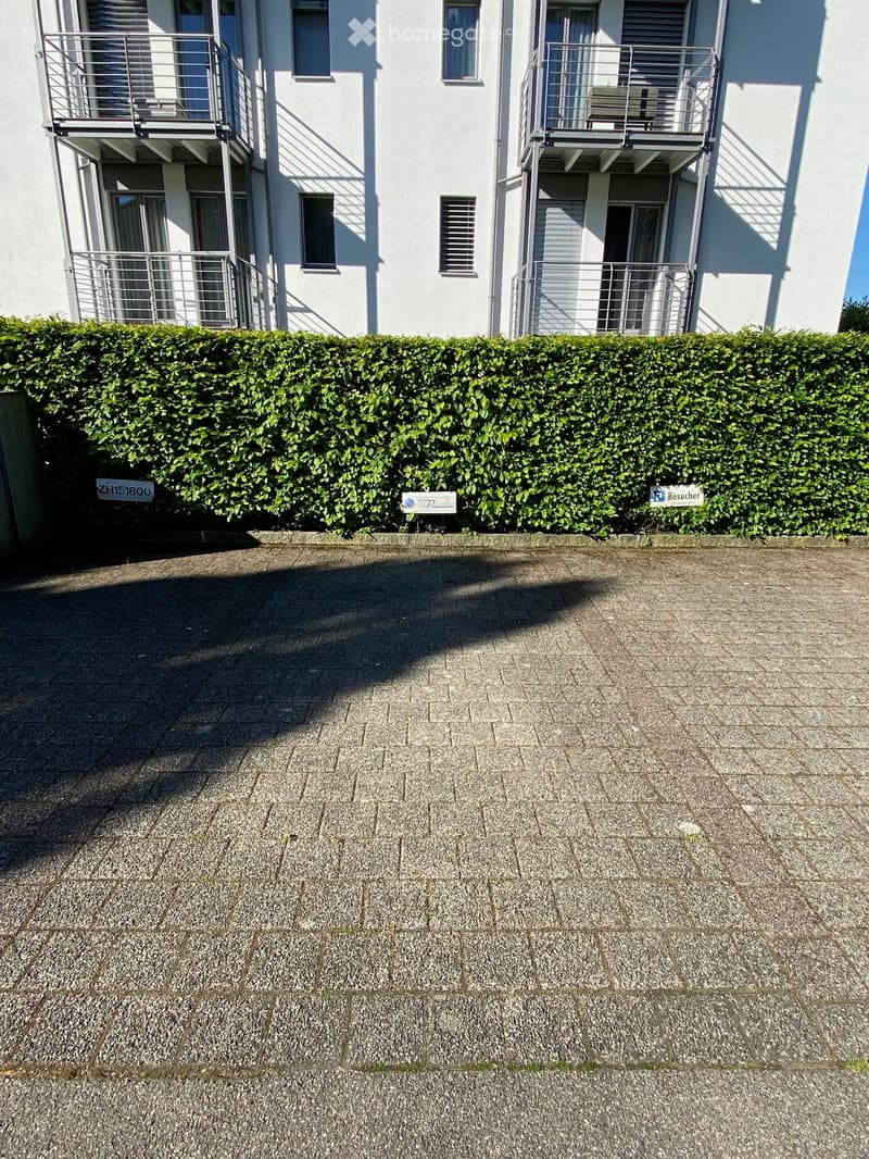A paved outdoor area in front of a white multi-story building with balconies, featuring a neatly trimmed hedge and signs for visitors and real estate.