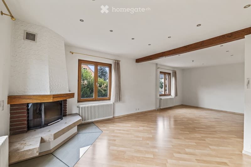 Empty living room with wooden flooring, white walls, exposed wooden beams, fireplace, large windows with curtains, and radiators.
