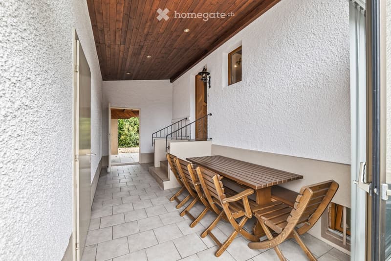 Balcony with wooden table and chairs, tiled floor, wooden ceiling, white walls, open doorway leading to another area.