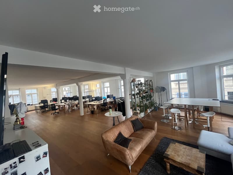 A spacious, modern office or lounge area with wooden flooring, large windows, a brown leather sofa, coffee table, several chairs, a large screen, and multiple laptops on tables.